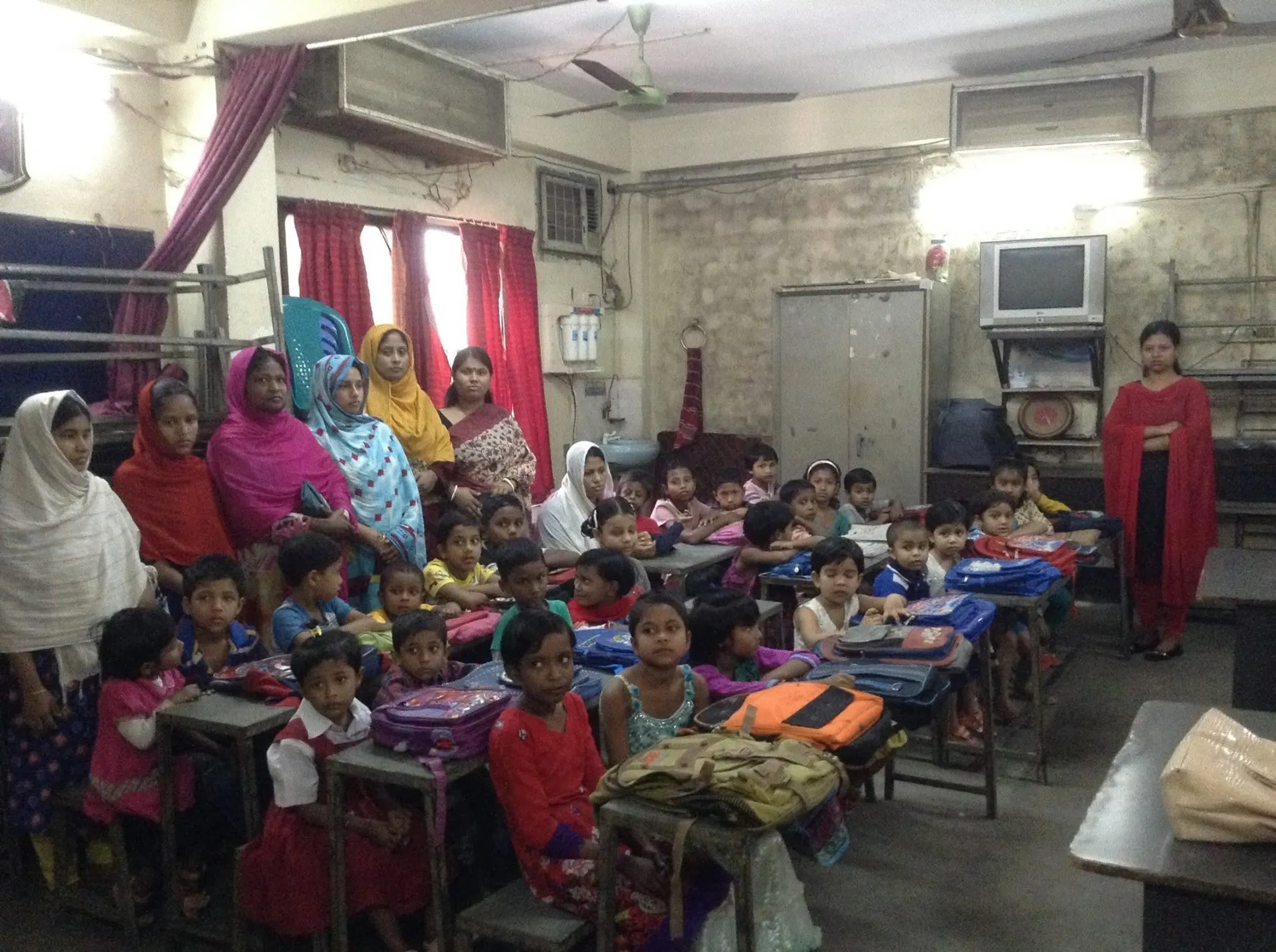 Young students at their desks with new school bags while their mothers stand at the back of the classroom