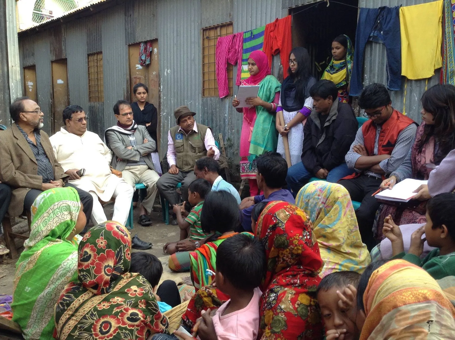 Women, children and community leaders seated together at an outdoor meeting in a slum courtyard