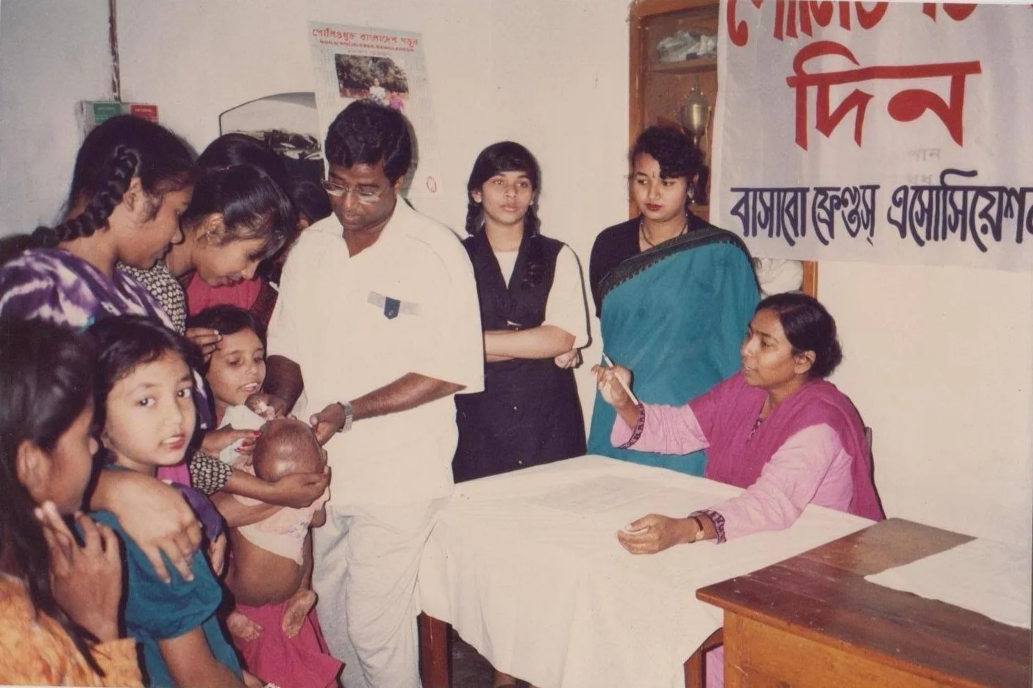 A volunteer gives a baby vaccine drops while a woman keeps the register at a BFA immunization day, in an old printed photograph